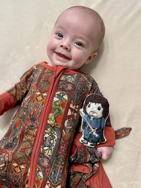 Smiling baby lying in the Lord of the Rings Stained Glass zip romper with the Aragorn lovey beside them