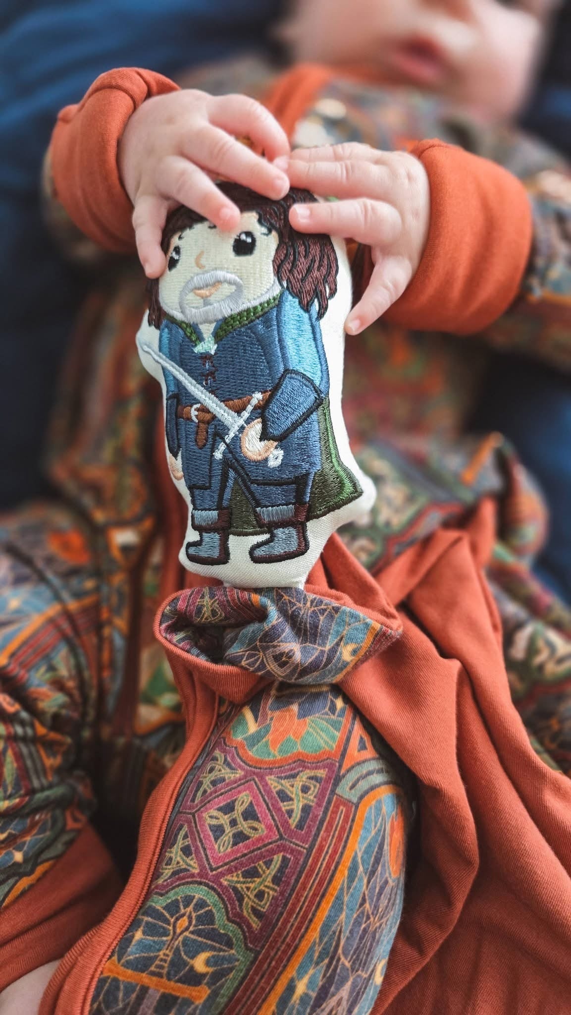 Baby holding the embroidered Aragorn lovey, knotted corner tied, over the matching Lord of the Rings Stained Glass zip romper