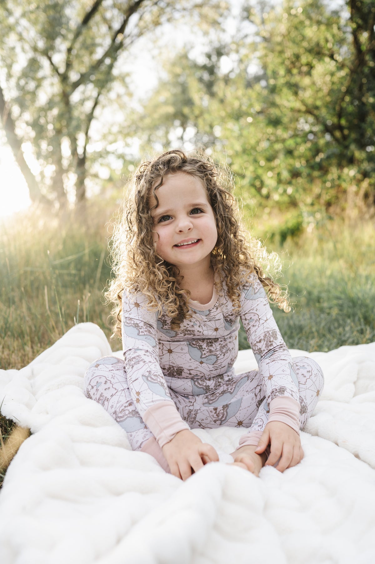 Toddler girl smiling on a blanket in the grass, wearing the Tiny Maps LOTR two-piece pajama set